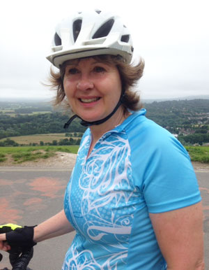 Lorraine Barratt in a white cycling helmet and blue jersey, smiling, stopped at the top of a hill with the Sussex countryside behind her